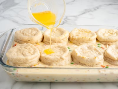 A glass baking dish filled with creamy Chicken Pot Pie Casserole, topped with golden biscuits; melted butter is being poured over the biscuits from a small glass bowl.
