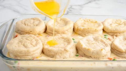A glass baking dish filled with creamy Chicken Pot Pie Casserole, topped with golden biscuits; melted butter is being poured over the biscuits from a small glass bowl.