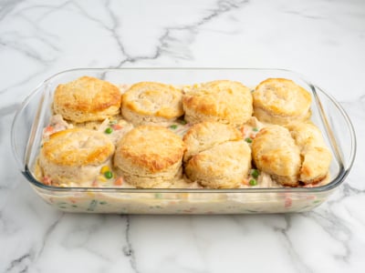 A glass baking dish filled with creamy Chicken Pot Pie Casserole, topped with golden-brown biscuits, showing mixed vegetables and chicken in the filling, on a white marble surface.