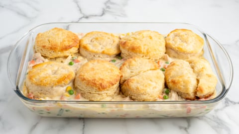 A glass baking dish filled with creamy Chicken Pot Pie Casserole, topped with golden-brown biscuits, showing mixed vegetables and chicken in the filling, on a white marble surface.