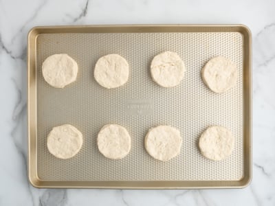 A gold baking sheet with eight evenly spaced, round raw biscuit dough pieces—perfect for topping a Chicken Pot Pie Casserole—arranged in two rows on a white marble surface.