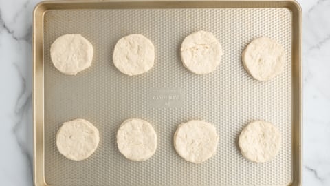 A gold baking sheet with eight evenly spaced, round raw biscuit dough pieces—perfect for topping a Chicken Pot Pie Casserole—arranged in two rows on a white marble surface.