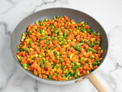 A frying pan with a mix of diced carrots, green beans, peas, and corn—classic Chicken Pot Pie Casserole vegetables—sits on a white marble surface. The veggies are colorful and appear freshly cooked.