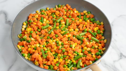 A frying pan with a mix of diced carrots, green beans, peas, and corn—classic Chicken Pot Pie Casserole vegetables—sits on a white marble surface. The veggies are colorful and appear freshly cooked.