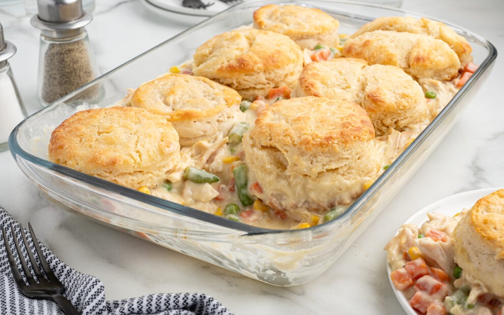 A glass baking dish filled with creamy Chicken Pot Pie Casserole, topped with golden-brown biscuits. A serving is plated nearby, with a fork and striped napkin beside it. Pepper and salt shakers are in the background.