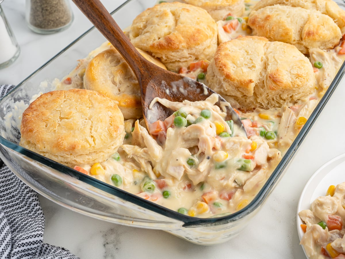 A glass baking dish filled with creamy Chicken Pot Pie Casserole, topped with golden biscuits. A wooden spoon scoops out a portion, revealing peas, carrots, corn, and chicken in a rich sauce.
