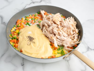 A skillet containing mixed vegetables, shredded cooked chicken, and a creamy sauce with black pepper—like the filling of a Chicken Pot Pie Casserole—all arranged separately and ready to be mixed together.
