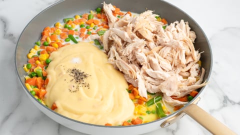 A skillet containing mixed vegetables, shredded cooked chicken, and a creamy sauce with black pepper—like the filling of a Chicken Pot Pie Casserole—all arranged separately and ready to be mixed together.