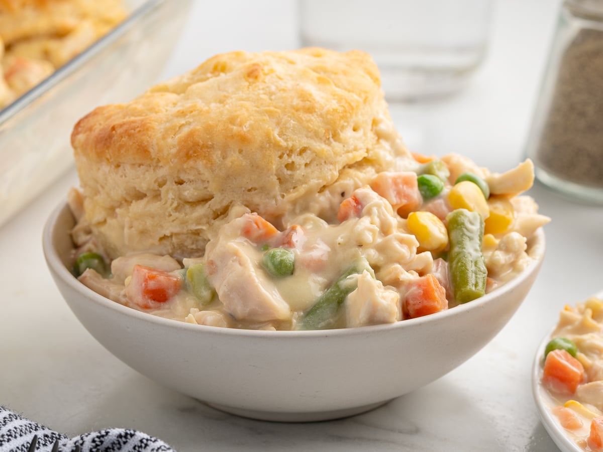 A bowl of creamy Chicken Pot Pie Casserole filling topped with a golden, flaky biscuit. The hearty filling contains carrots, peas, corn, and green beans. A baking dish and pepper shaker sit in the background.
