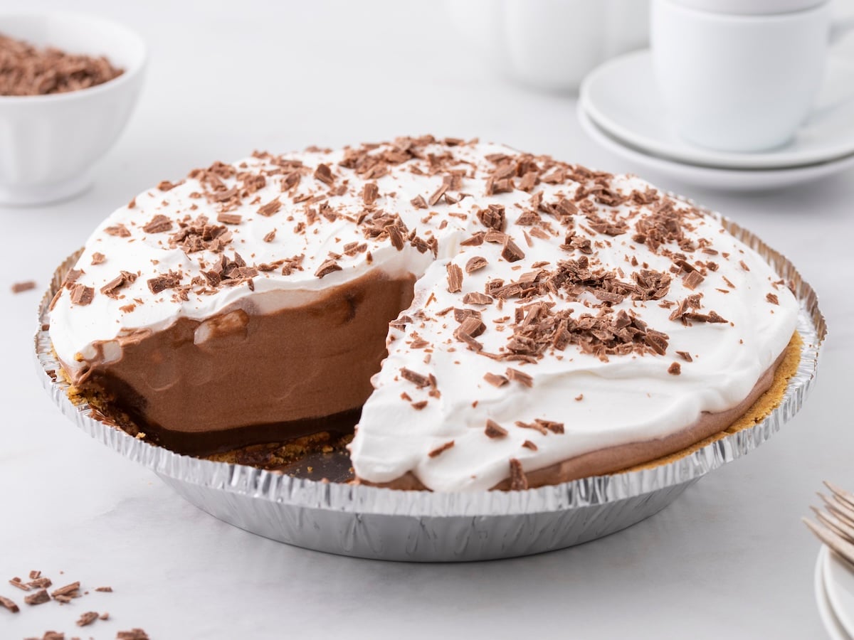 A Chocolate Pudding Pie with a slice missing, topped with whipped cream and chocolate shavings, sits in a foil tin on a white surface. This classic Chocolate Pudding Pie Recipe is served with cups, saucers, and a bowl in the background.
