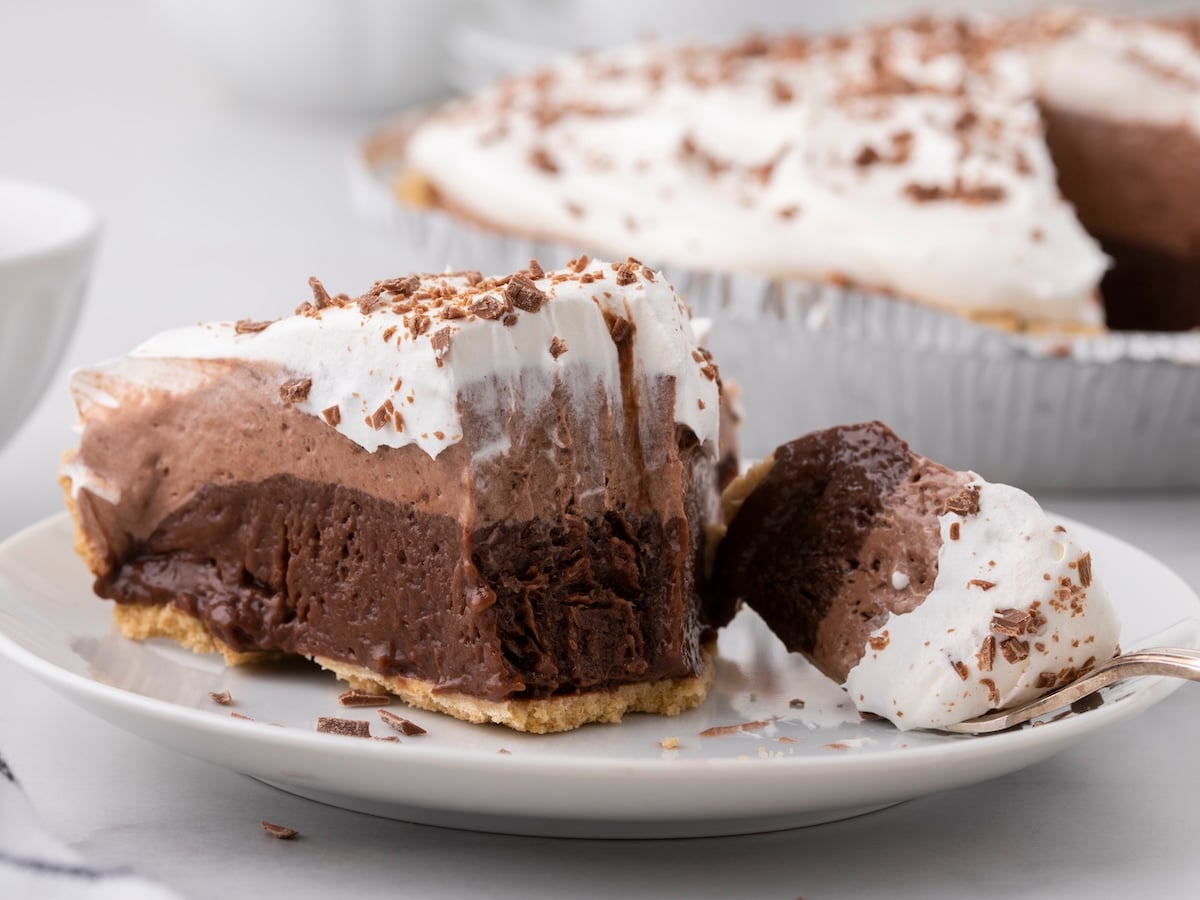 A slice of layered Chocolate Pudding Pie with whipped cream and chocolate shavings on top is served on a plate, with a bite taken out and placed on a fork. The whole pie is visible in the background, tempting you to try this Chocolate Pudding Pie Recipe.