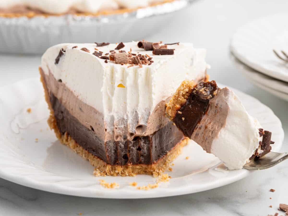 A slice of layered Chocolate Pudding Pie with a graham cracker crust, topped with whipped cream and chocolate shavings, sits on a white plate. A fork holds a bite of the pie in the foreground, tempting you to try this irresistible Chocolate Pudding Pie Recipe.