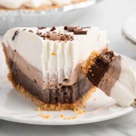 A slice of layered Chocolate Pudding Pie with a graham cracker crust, topped with whipped cream and chocolate shavings, sits on a white plate. A fork holds a bite of the pie in the foreground, tempting you to try this irresistible Chocolate Pudding Pie Recipe.