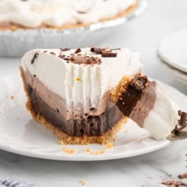 A slice of layered Chocolate Pudding Pie with a graham cracker crust, topped with whipped cream and chocolate shavings, sits on a white plate with a forkful taken out.