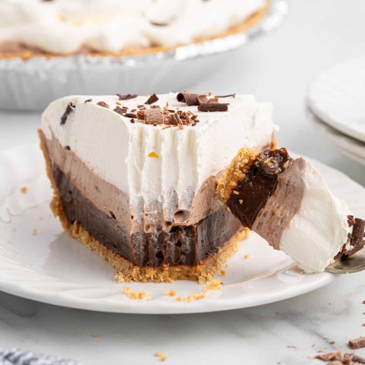 A slice of layered Chocolate Pudding Pie with a graham cracker crust, topped with whipped cream and chocolate shavings, sits on a white plate with a forkful taken out.