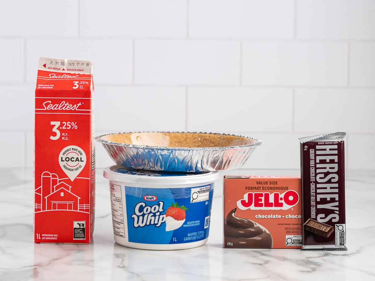 On a white countertop with a tiled wall background, ingredients for a Chocolate Pudding Pie are arranged: a carton of milk, tub of Cool Whip, chocolate Jello pudding box, Hersheyโs chocolate bar, and a graham pie crustโeverything you need to start your favorite Chocolate Pudding Pie Recipe.
