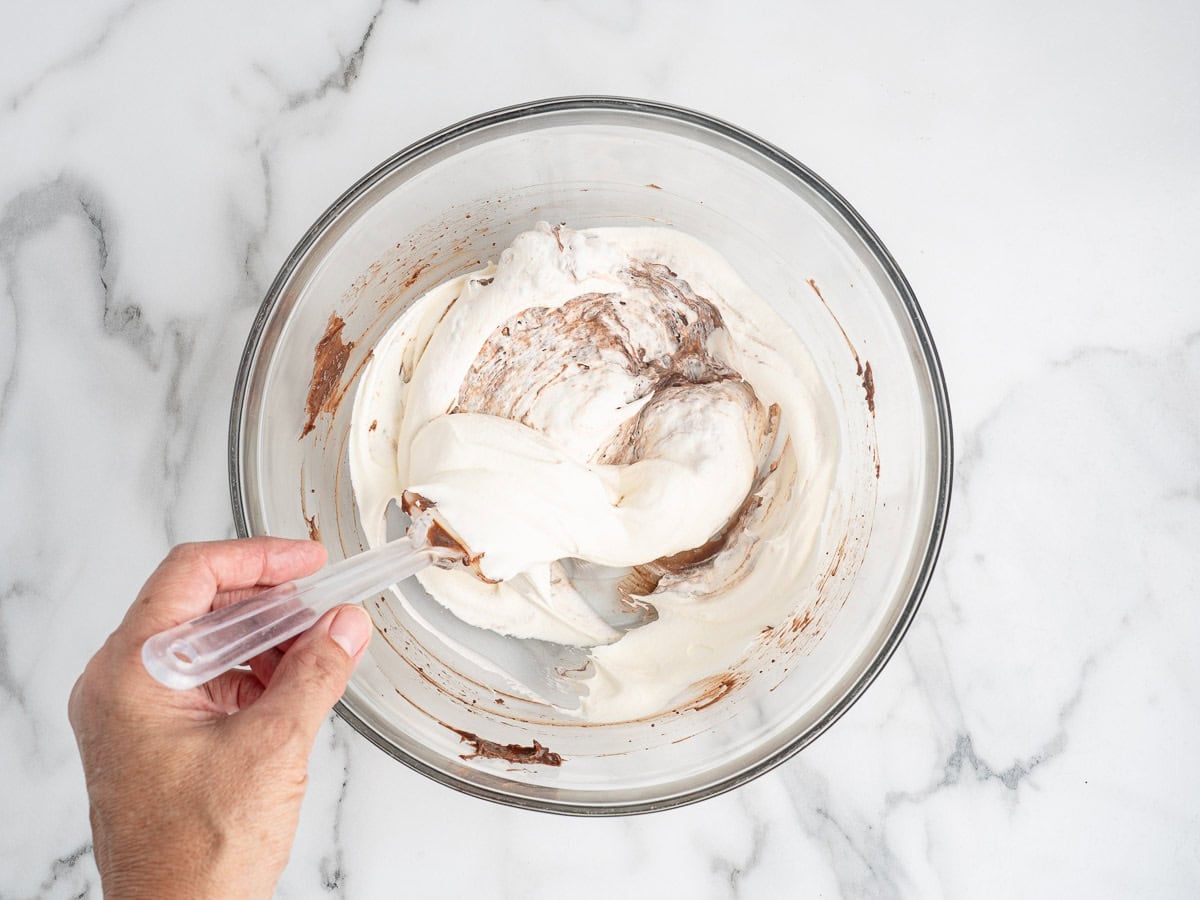 A hand using a spatula to fold whipped cream into chocolate mixture in a glass bowl on a marble countertop, preparing the creamy filling for a classic Chocolate Pudding Pie Recipe.