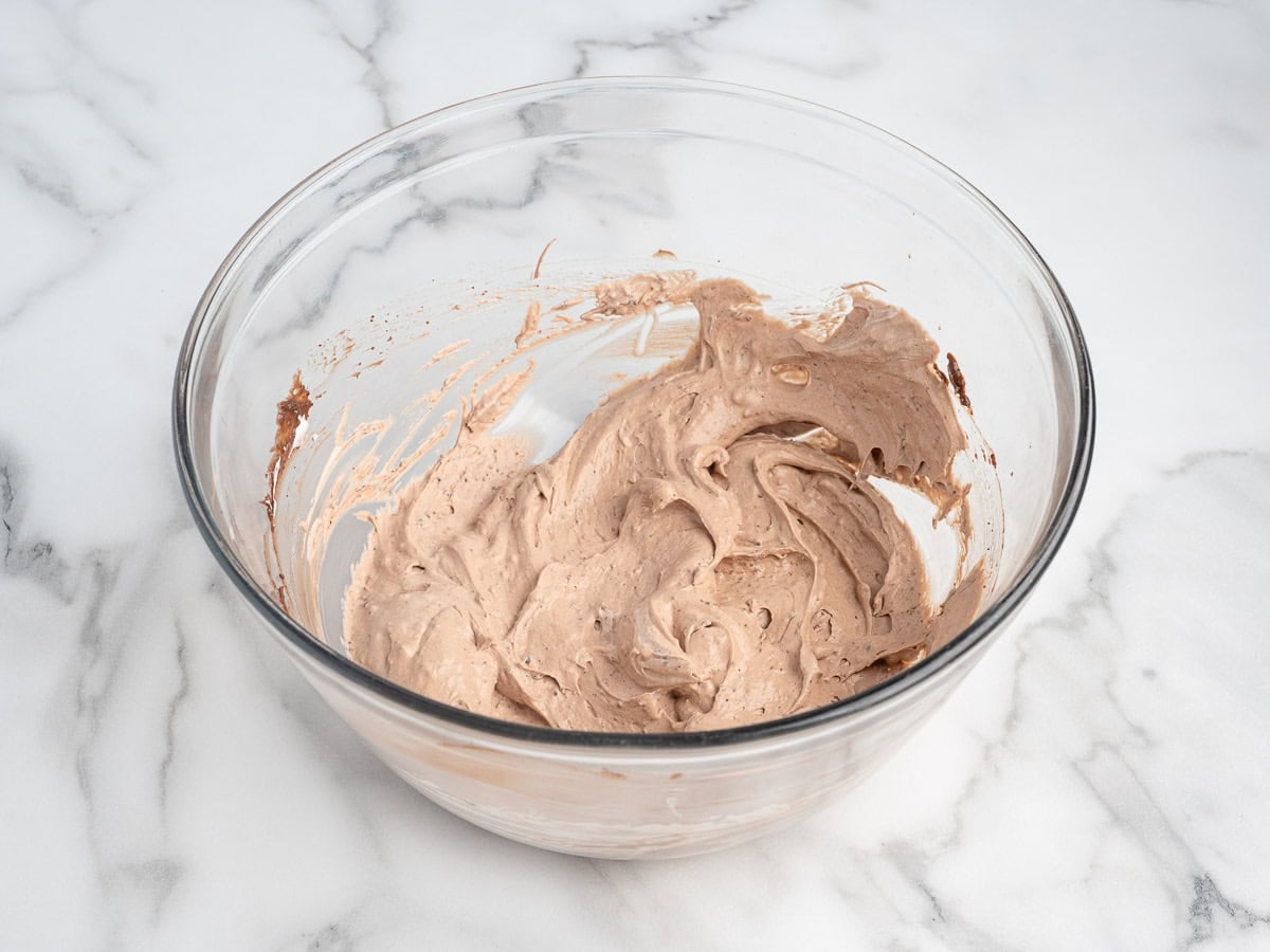 A glass bowl on a marble surface containing whipped chocolate mousse or frosting, with swirls and soft peaks visible in the light brown mixtureโperfect as a decadent topping or filling for your favorite Chocolate Pudding Pie Recipe.