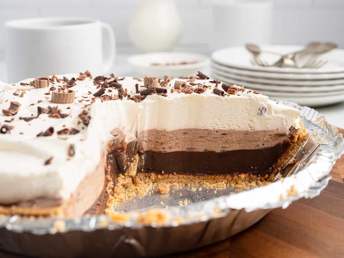 A classic Chocolate Pudding Pie recipe featuring a chocolate cream pie with a graham cracker crust and whipped cream topping, sliced to show its luscious layered chocolate filling and garnished with chocolate shavings. Plates, forks, and a cup are in the background.