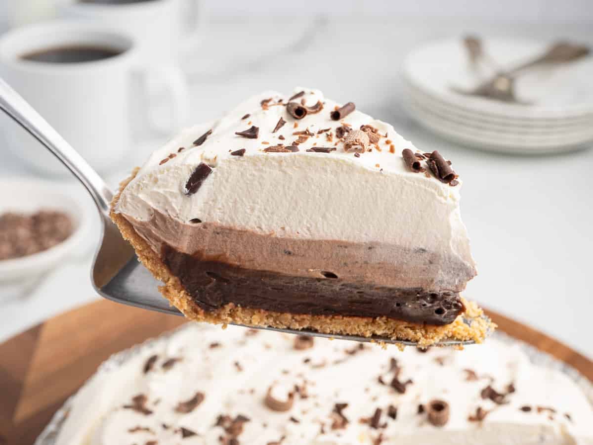 A close-up of a pie slice with layers of chocolate and whipped cream, topped with chocolate shavings, held above the whole Chocolate Pudding Pie on a cake server. Plates, forks, and coffee cups are in the background, perfectly capturing the irresistible appeal of this classic Chocolate Pudding Pie Recipe.