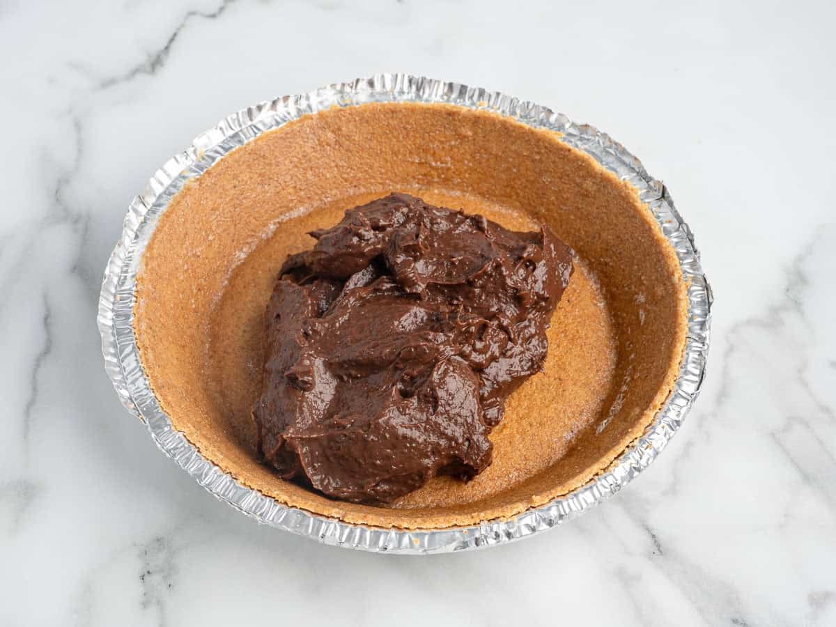 A foil pie tin with a graham cracker crust is filled with a large scoop of rich chocolate pie filling, showcasing a classic Chocolate Pudding Pie Recipe on a white marble surface.