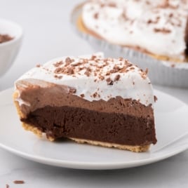 A slice of layered chocolate cream pie with a chocolate base, creamy middle, and whipped topping—just like a classic Chocolate Pudding Pie Recipe—garnished with chocolate shavings, sits on a white plate. The remaining pie is in the background.