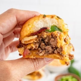 A hand holds a half-eaten Cowboy Slider with ground beef, bacon, cheese, jalapeño, and onions, showing the juicy filling and savory toppings.