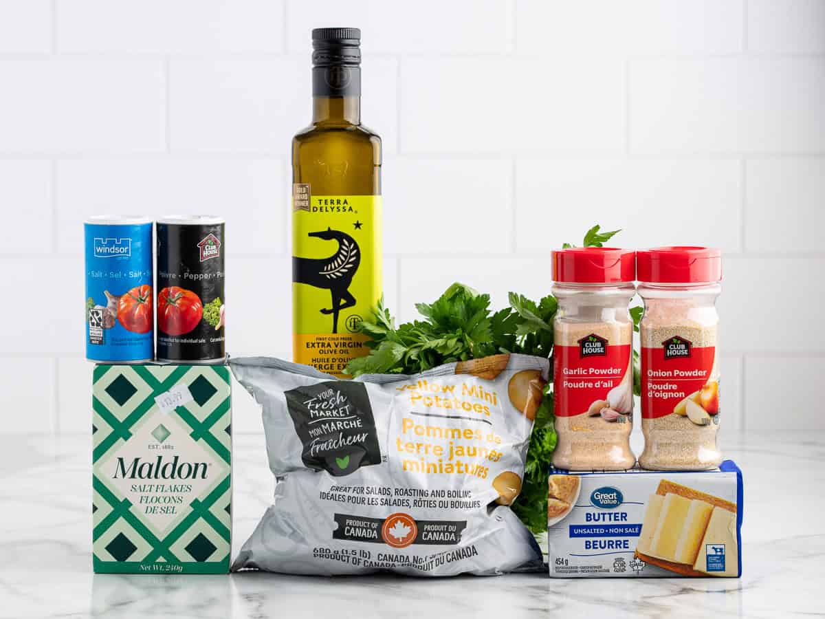 A variety of ingredients on a kitchen counter, including spices, olive oil, parsley, a bag of yellow mini potatoes perfect for making crispy smashed potatoes, butter, and canned tomatoes, against a white tiled background.