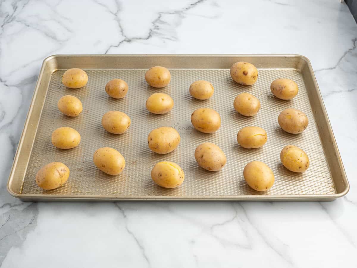 A baking sheet with 20 small, unpeeled yellow potatoes evenly spaced on a white marble countertop, ready to be transformed into delicious Crispy Smashed Potatoes.