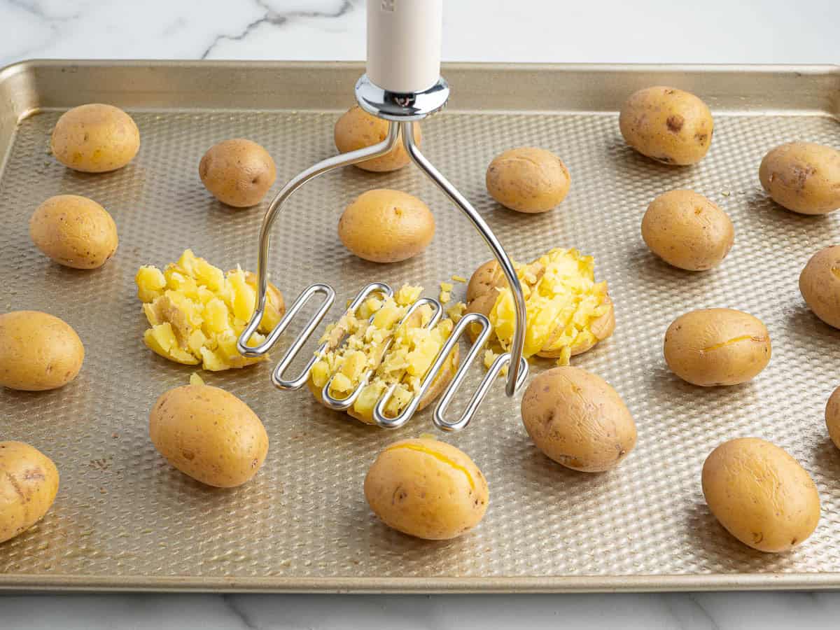 A potato masher is pressing down on small yellow potatoes on a baking sheet, partially smashing one potato while others remain whole, creating the perfect base for Crispy Smashed Potatoes.