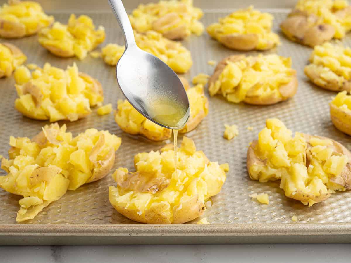 A silver spoon drizzles oil over smashed yellow potatoes arranged on a baking sheet, ready to be roasted into perfectly crispy smashed potatoes.