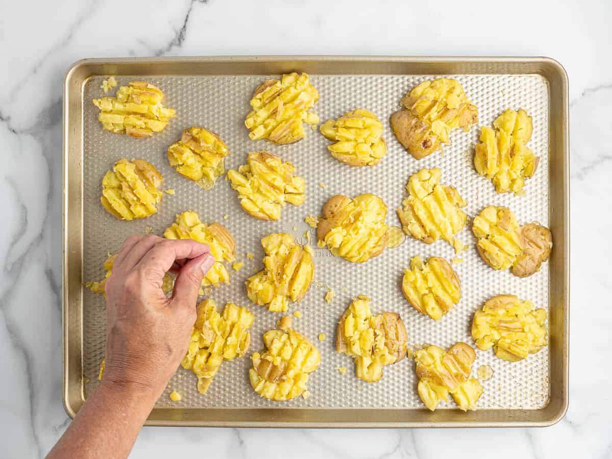 A hand sprinkles seasoning on crispy smashed potatoes arranged on a baking sheet, ready for roasting. The tray sits on a white marble surface.
