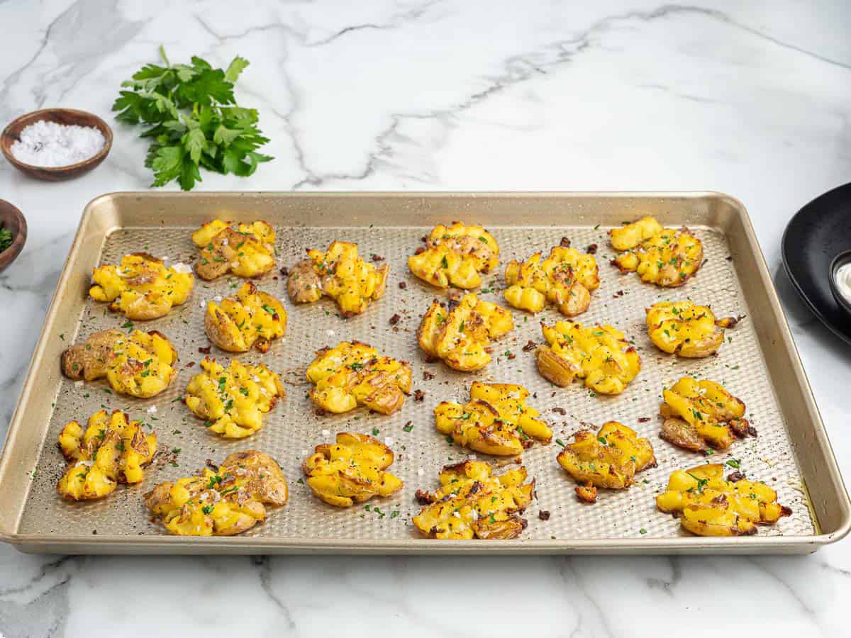 Golden crispy smashed potatoes garnished with herbs on a baking sheet, with small bowls of salt and fresh parsley on the side, set on a white marble countertop.