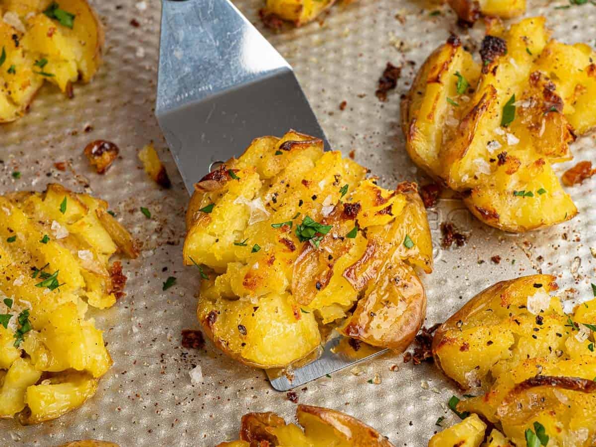 A metal spatula lifts a golden, crispy smashed potato garnished with herbs and salt from a baking tray filled with more perfectly baked Crispy Smashed Potatoes.