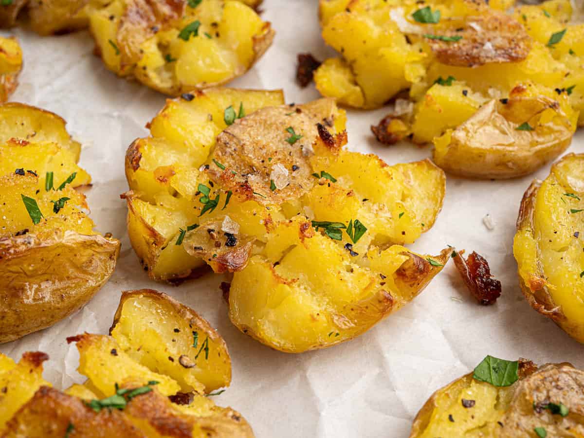 Golden crispy smashed potatoes with perfectly crunchy edges, sprinkled with chopped herbs, coarse salt, and black pepper, beautifully arranged on parchment paper.