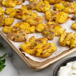 A baking sheet holds crispy smashed potatoes, their golden edges perfectly browned and garnished with herbs. A bowl of sour cream and fresh parsley are nearby on a white countertop, ready to complement the irresistible Crispy Smashed Potatoes.