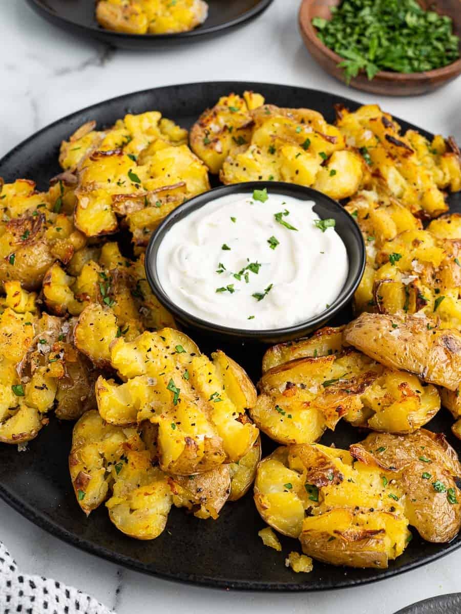 A black plate of crispy smashed potatoes garnished with chopped herbs, surrounding a small bowl of white dipping sauce, is beautifully arranged on a marble surface—perfect for anyone craving restaurant-style Crispy Smashed Potatoes at home.