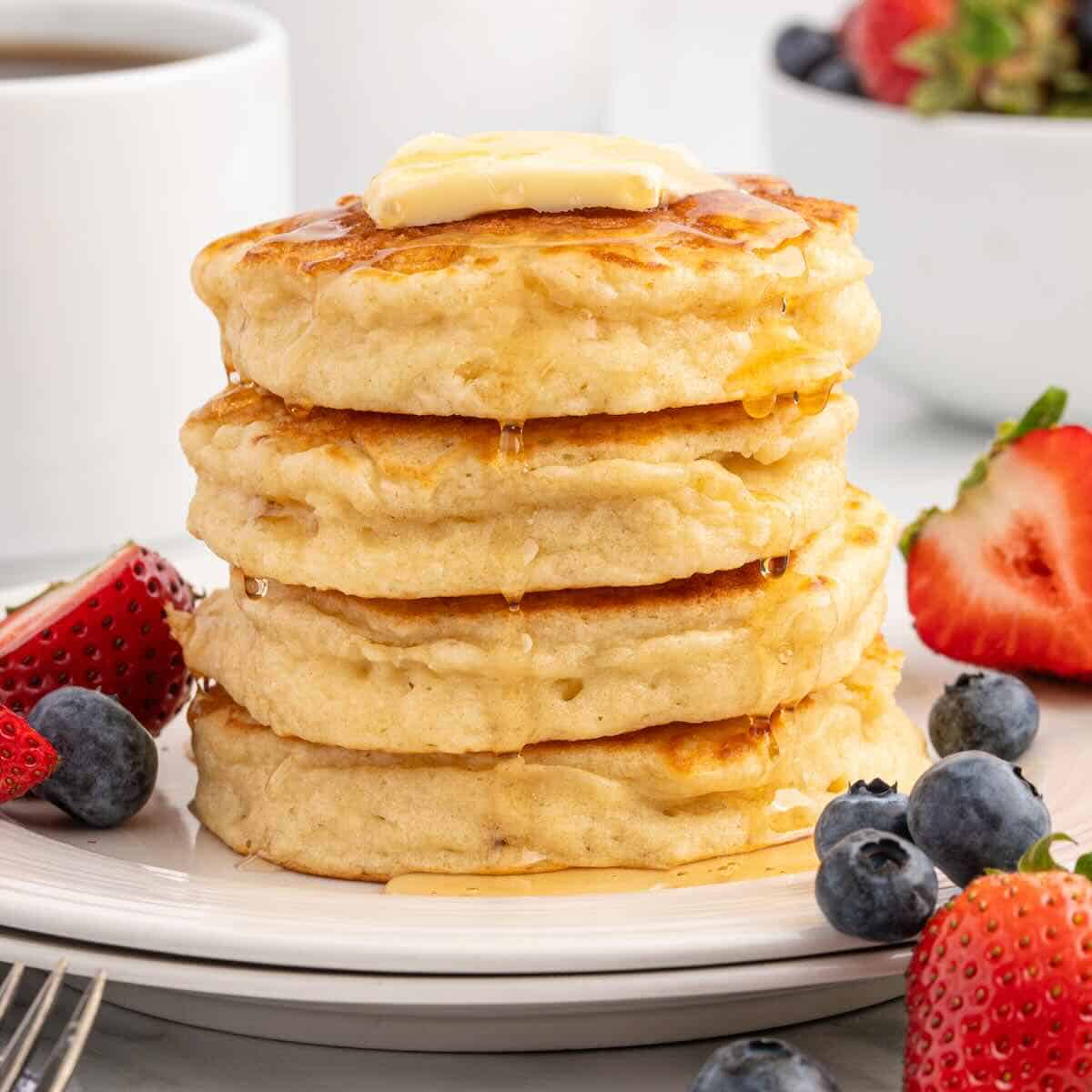 A stack of four pancakes from our favorite Fluffy Pancake Recipe is topped with butter and syrup, surrounded by fresh strawberries and blueberries on a white plate.