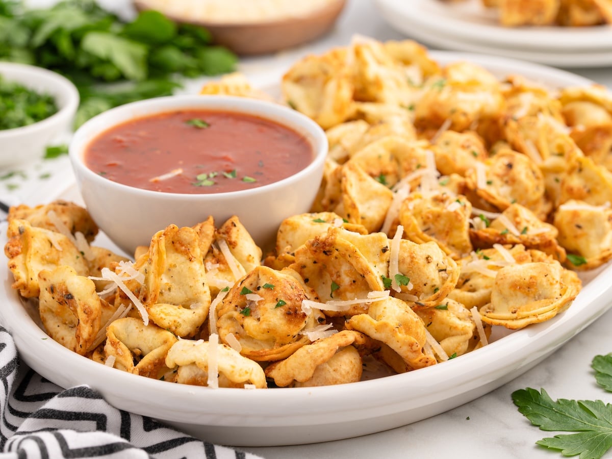 A white plate filled with crispy Air Fryer Tortellini, garnished with shredded cheese and herbs, is served with a bowl of red marinara sauce for dipping. Fresh parsley and a striped napkin are in the background.