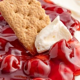 A side view of a dish filled with cheesecake dip and topped with glossy cherry pie filling. A graham cracker is being dipped into the dessert.