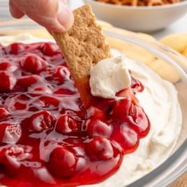 A graham cracker being dipped into a bowl of cheesecake dip topped with cherry pie filling, surrounded by additional crackers and pretzels in the background.