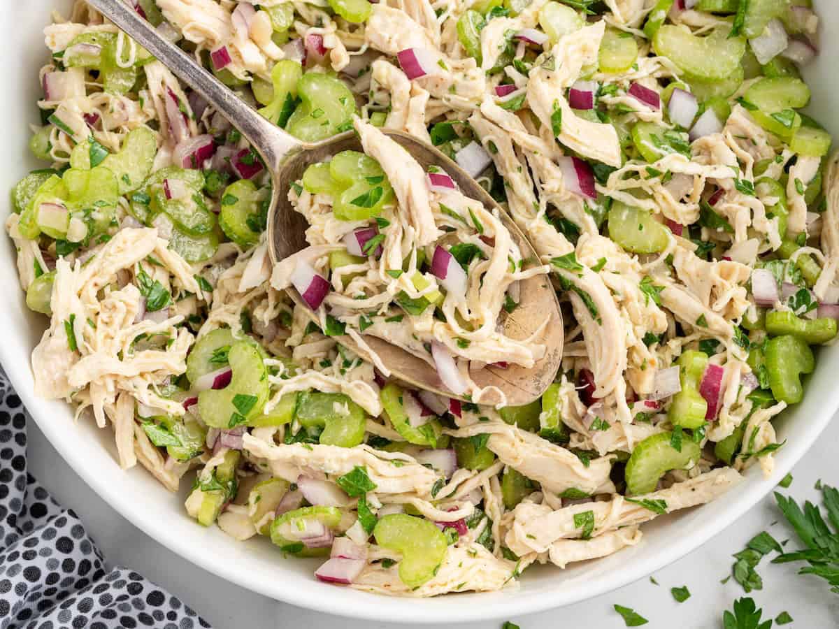 A bowl of chicken salad without mayo features shredded chicken, sliced celery, chopped red onion, and parsley, all mixed with a light dressing. A large spoon is scooping out a portion.
