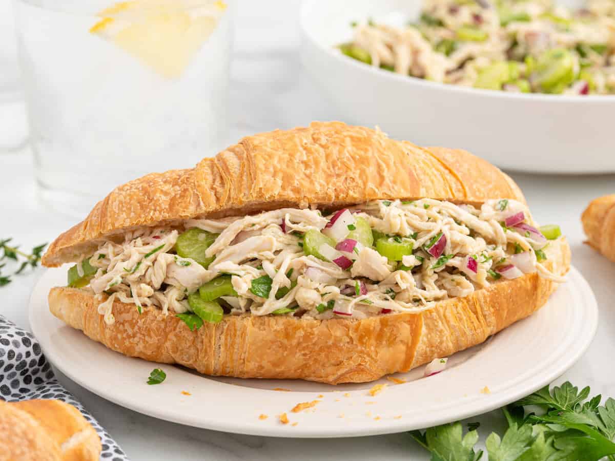A croissant sandwich filled with chicken salad without mayo, celery, and red onion sits on a white plate. A glass of water with lemon and a bowl of chicken salad are in the background. Parsley garnish is visible on the side.