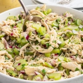 A white bowl filled with shredded chicken, chopped celery, red onion, and fresh herbs is being mixed with a large spoon, creating a flavorful Chicken Salad Without Mayo. Plates are stacked in the background.