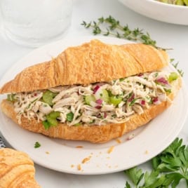 A croissant sandwich filled with shredded chicken, chopped celery, red onion, and herbs sits on a white plate with fresh parsley and herbs in the background—perfect for those seeking a flavorful chicken salad without mayo.