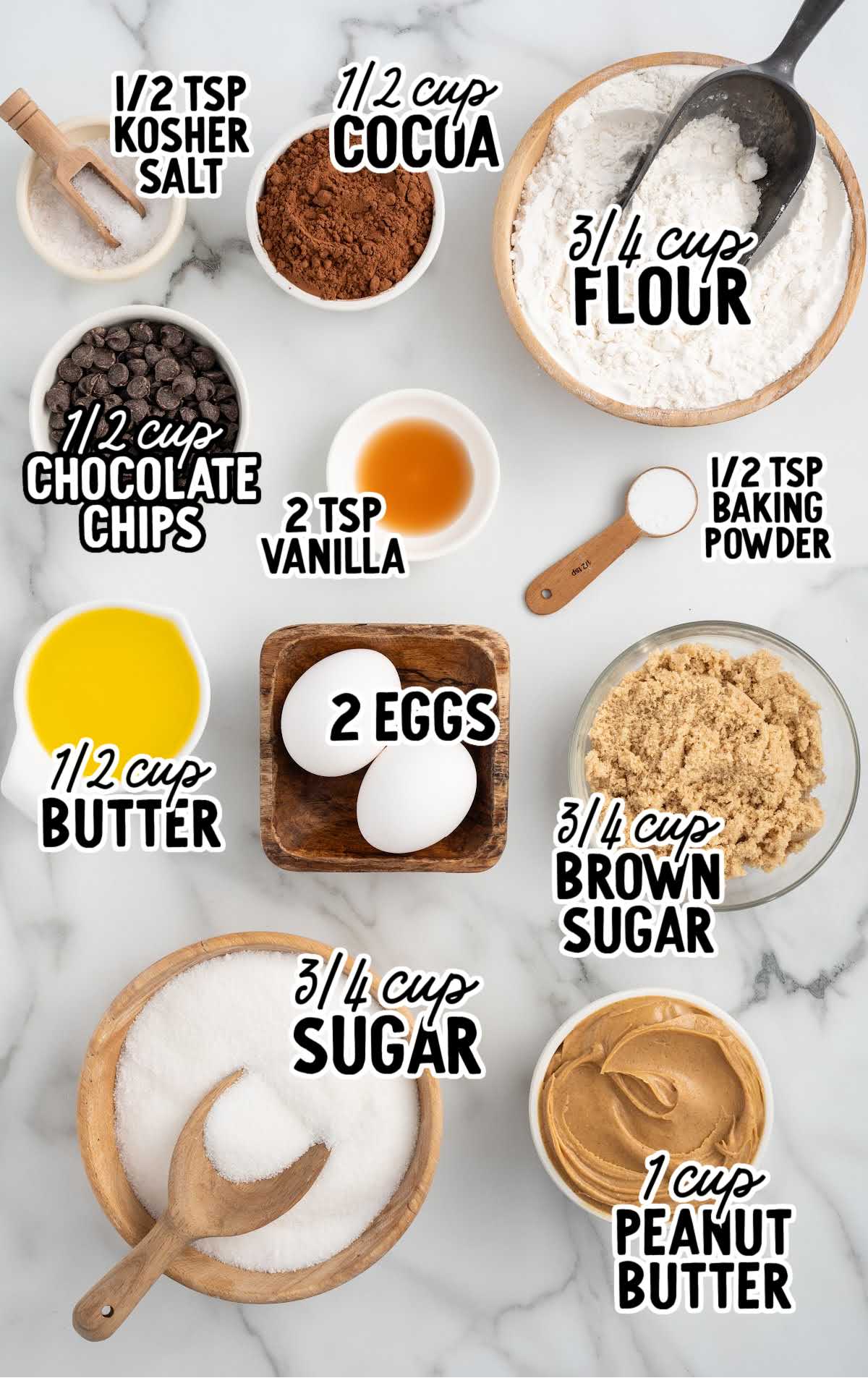 A flat lay of ingredients for chocolate peanut butter brownies, including flour, cocoa powder, sugar, butter, eggs, peanut butter, chocolate chips, and vanilla, all neatly labeled on a marble background.