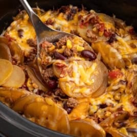 A close-up of a Crock Pot Cowboy Casserole filled with cheesy potatoes, ground beef, beans, and diced tomatoes, with a serving spoon lifting a portion.