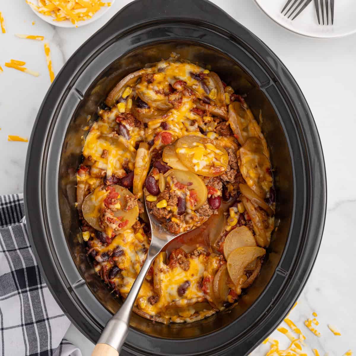 Top-down view of a black slow cooker filled with Crock Pot Cowboy Casserole—a cheesy potato and ground beef casserole loaded with kidney beans and corn. A serving spoon lifts some out, showing the hearty layers. Shredded cheddar and a napkin rest on the white marble surface nearby.
