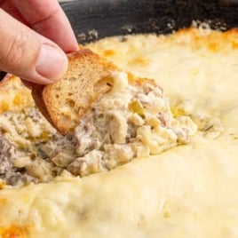 A skillet filled with freshly baked Philly cheesesteak dip with a golden-brown top, paired with a bowl of toasted bread slices.