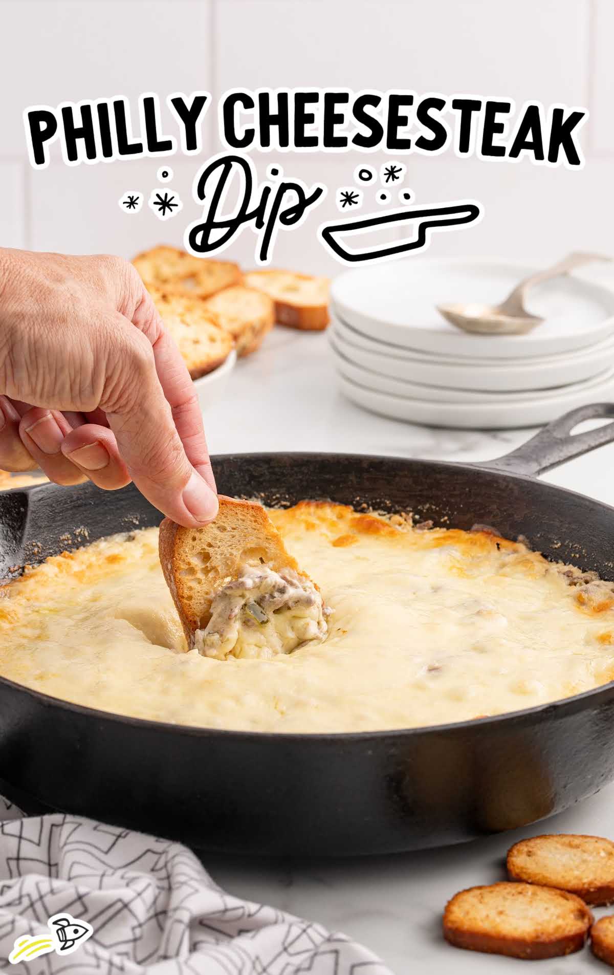 A hand dipping a slice of toasted bread into a skillet of gooey Philly cheesesteak dip, emphasizing the rich, cheesy texture.